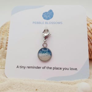 May include: A silver charm with a round pendant depicting a beach scene with blue and white waves over sand. The charm has a lobster clasp. The card behind the charm says "Pebble Blossoms" and "A tiny reminder of the place you love."