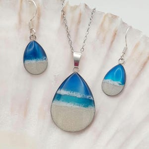 May include: A jewellery set consisting of a teardrop-shaped pendant on a silver chain and two matching teardrop-shaped earrings. Each piece features a gradient design transitioning from deep blue to light blue and a textured sandy beige base.