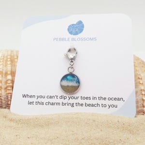 Peut inclure: Un charm circulaire argenté représentant une scène de plage miniature avec du sable beige et des vagues bleues à l'écume blanche. Le charm est fixé à un fermoir mousqueton argenté sur une carte de présentation blanche PEBBLE BLOSSOMS.