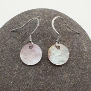 May include: A pair of drop earrings featuring circular, iridescent mother-of-pearl discs suspended from silver-toned metal fishhook wires. The discs exhibit a smooth, polished surface with subtle variations in white and soft pinkish hues.