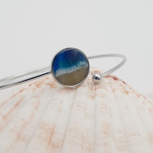May include: A silver-toned metal cuff bracelet featuring a circular pendant. The pendant contains a resin inlay depicting a coastal scene with a deep blue ocean, a white foamy wave, and a textured tan sandy beach at the bottom of the circle.