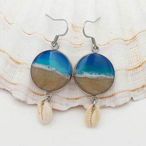 May include: A pair of circular drop earrings featuring a resin design of a sandy beach meeting blue ocean waves with white foam. Each earring is suspended from a silver-toned metal hook and includes a small, cream-coloured cowrie shell charm.