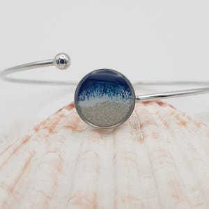 May include: A silver cuff bracelet with a circular ocean-themed charm. The charm depicts a blue and white wave design above a sandy base. The bracelet has a minimalist, open design with a small ball detail at one end.