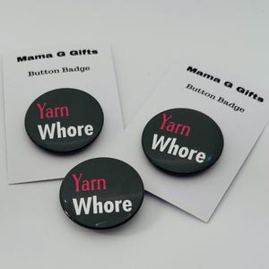 以下が含まれることがあります： 白とピンクの文字で「Yarn Whore」と書かれた3つの黒いボタンバッジ。バッジは「Mama G Gifts」と「Button Badge」と書かれた白いカードにあります。