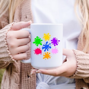 May include: White ceramic mug with a colorful paint splatter design. The mug has a handle and is held by a person's hands.