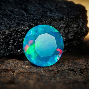 May include: A round, faceted, blue opal gemstone with green and pink flashes of color. The opal is sitting on a dark brown, textured surface.
