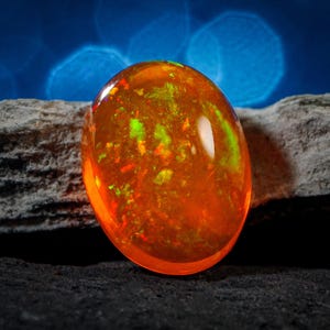 Könnte beinhalten: Ein leuchtend orangefarbener Opal-Edelstein mit grünen und roten Farbschimmern. Der Opal ist oval geformt und liegt auf einem grauen Stein.