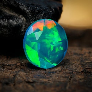 May include: A round, faceted opal gemstone with a blue-green hue and iridescent flashes of color. The opal is resting on a dark, textured surface.