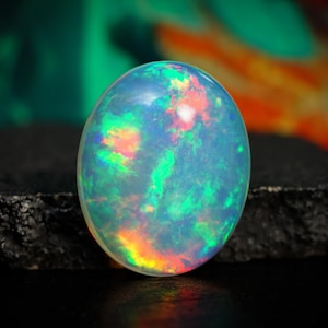 May include: A polished, oval opal gemstone. The opal displays a vibrant play-of-color, with flashes of green, blue, orange, and red. The gemstone is set against a dark, textured surface, with a blurred background of green and orange.