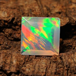 Puede incluir: Una piedra preciosa de ópalo de forma cuadrada, transparente, con un arcoíris de colores, incluyendo verde, rojo y naranja, sobre una superficie marrón texturizada.