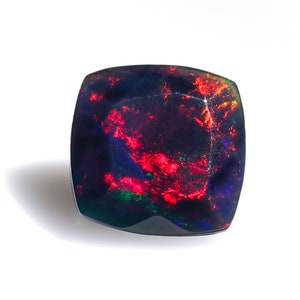 Puede incluir: Una piedra preciosa de ópalo negro oscuro con destellos rojos, verdes y azules. La piedra preciosa está tallada en forma cuadrada con bordes redondeados.