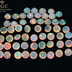 May include: A collection of round, faceted opals displaying iridescent flashes of orange, green, and blue against a translucent white base. The stones are arranged in a grid pattern. The top left corner features the text HGC House of Gems Creations Since 1985.