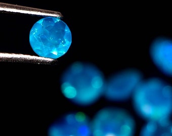 May include: A close-up of a bright blue gemstone held by tweezers. The gemstone is faceted and has a star-like pattern. Other blue gemstones are out of focus in the background.