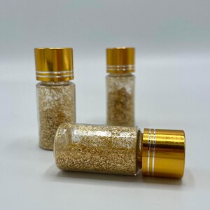 Könnte beinhalten: Drei kleine Glasflaschen mit goldfarbenen Deckeln. Die Flaschen sind mit einer goldfarbenen, glitzerartigen Substanz gefüllt. Eine Flasche liegt auf der Seite, während die anderen beiden aufrecht stehen. Der Hintergrund ist einfarbig hellgrau.