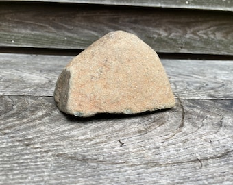 May include: A light brown, irregularly shaped rock with a smooth, rounded surface. The rock is sitting on a weathered wooden surface.