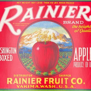 May include: Vintage Rainier brand apple crate label. The label features a large red apple in the center, with a mountain and orchard scene in the background. Text includes "Rainier Brand", "Washington Boxed", and "Apples Produce of U.S.A."