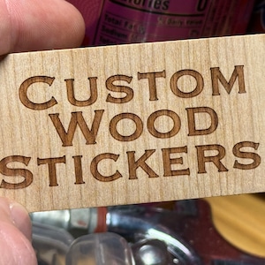 May include: A close-up of a wooden sign with the text "CUSTOM WOOD STICKERS!" engraved in brown lettering.