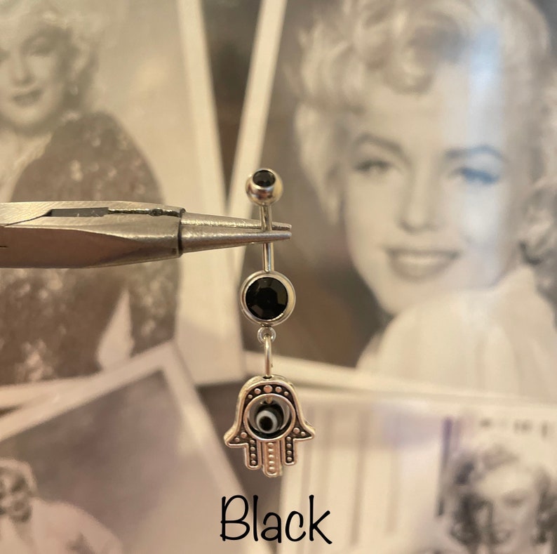 May include: A silver belly button ring with a black gem at the top and a larger black gem below. The charm is a silver Hamsa hand with an evil eye design. The word "Black" is written below the ring.