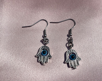 May include: A pair of silver earrings with a hamsa hand charm and a blue evil eye design.