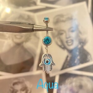 May include: A silver belly button ring with three aqua-colored gems and a dangling Hamsa hand charm. The Hamsa hand has an evil eye design in the center. The word "Aqua" is written in light blue script at the bottom.