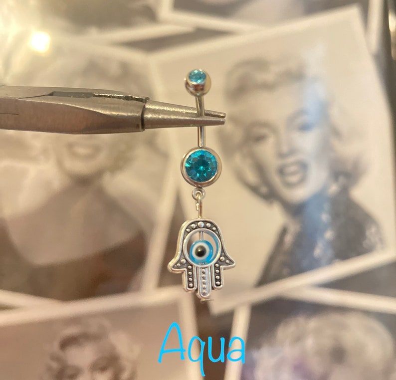May include: A silver belly button ring with three aqua-colored gems and a dangling Hamsa hand charm. The Hamsa hand has an evil eye design in the center. The word "Aqua" is written in light blue script at the bottom.