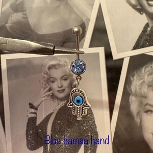 May include: A silver belly button ring with a light blue gem and a dangling Hamsa hand charm with a blue eye. The text "Blue hamsa hand" is at the bottom of the image.