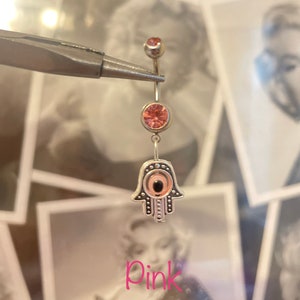 May include: A silver belly button ring with pink gemstones and a Hamsa hand charm. The Hamsa hand has an eye design in the center. The word "Pink" is written in pink script at the bottom of the image.