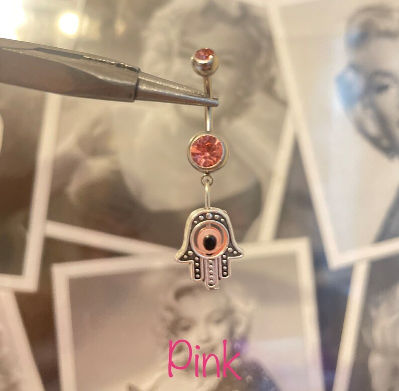 May include: A silver belly button ring with pink gemstones and a Hamsa hand charm. The Hamsa hand has an eye design in the center. The word "Pink" is written in pink script at the bottom of the image.
