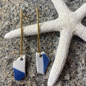 May include: A pair of gold earrings with blue and white ceramic pieces hanging from them. The earrings are on a white sandy surface with a starfish.