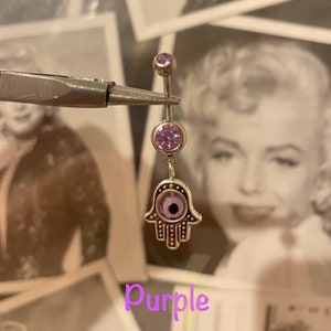 May include: A silver belly button ring with a dangling Hamsa hand charm. The ring features a purple gem at the top and a larger purple gem in the center. The Hamsa hand has a purple eye design. The word "Purple" is written in a purple font.