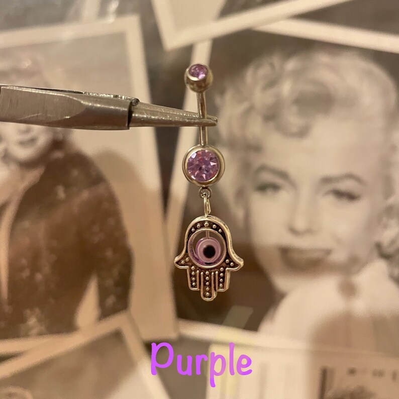 May include: A silver belly button ring with a dangling Hamsa hand charm. The ring features a purple gem at the top and a larger purple gem in the center. The Hamsa hand has a purple eye design. The word "Purple" is written in a purple font.