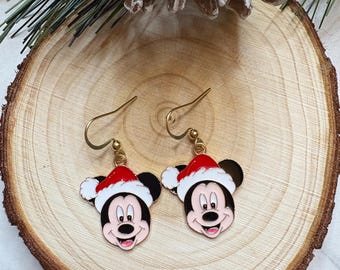 May include: A pair of gold-tone dangle earrings featuring Mickey Mouse wearing a Santa hat. The earrings have black ears, a red and white hat, and a smiling face. The earrings are displayed on a wooden slice.