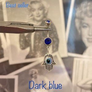 May include: A silver belly button ring with a dark blue gem at the top and a larger dark blue gem in the center. The charm is a silver Hamsa hand with an evil eye design. The text "Best seller" and "Dark blue" are visible.