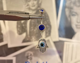 May include: A silver belly button ring with a dark blue gem at the top and a larger dark blue gem in the center. The charm is a silver Hamsa hand with an evil eye design. The text "Best seller" and "Dark blue" are visible.
