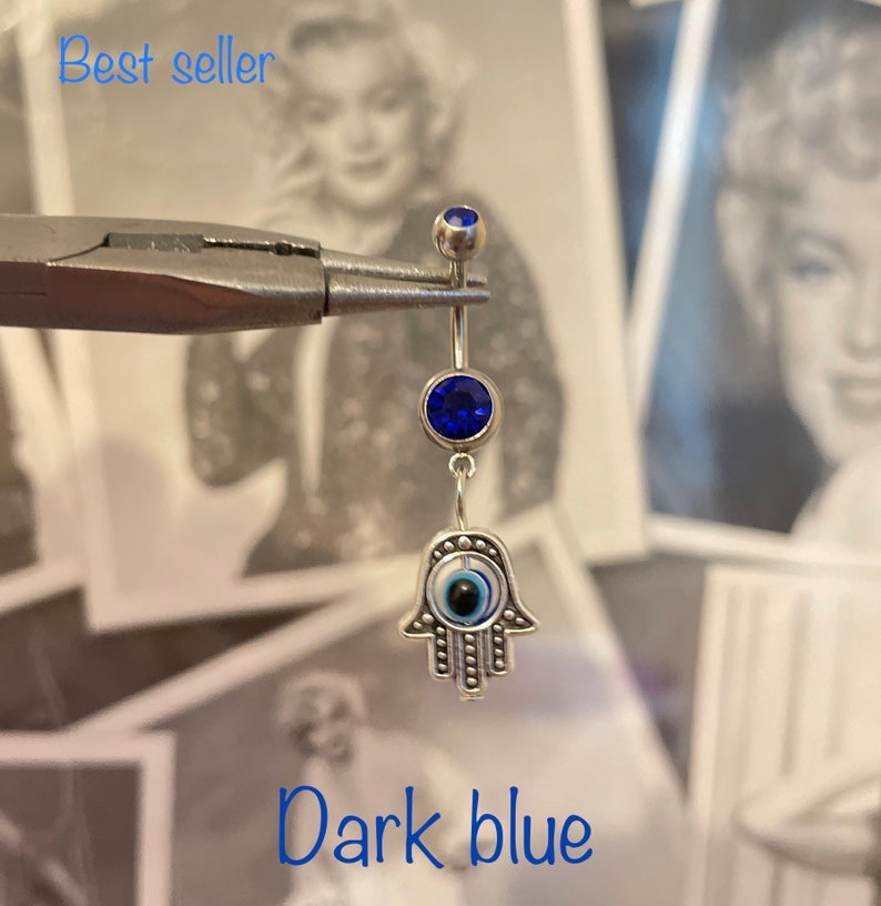 May include: A silver belly button ring with a dark blue gem at the top and a larger dark blue gem in the center. The charm is a silver Hamsa hand with an evil eye design. The text "Best seller" and "Dark blue" are visible.