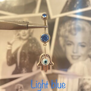 May include: A light blue belly button ring with a Hamsa hand charm. The ring features a small blue gem at the top and a larger blue gem below. The Hamsa hand charm has a white center with a blue eye design. The text "Light blue" is at the bottom.