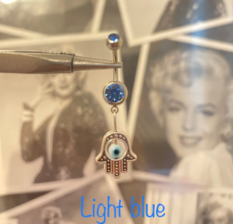 May include: A light blue belly button ring with a Hamsa hand charm. The ring features a small blue gem at the top and a larger blue gem below. The Hamsa hand charm has a white center with a blue eye design. The text "Light blue" is at the bottom.