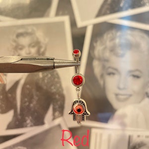 May include: A silver belly button ring with three red jewels and a Hamsa hand charm. The Hamsa hand has an eye in the center. The word "Red" is written in red script at the bottom of the image.