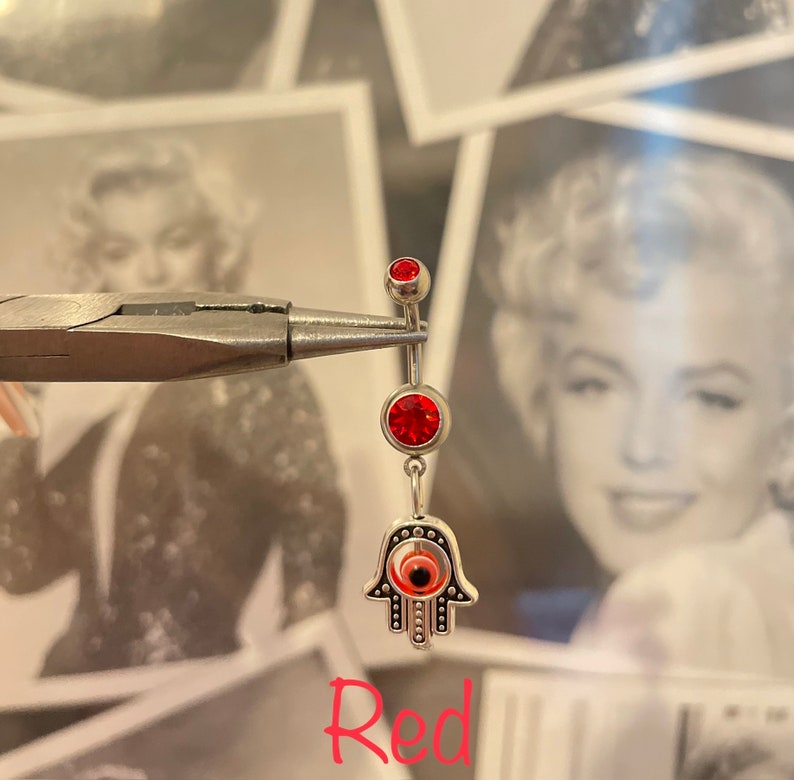 May include: A silver belly button ring with three red jewels and a Hamsa hand charm. The Hamsa hand has an eye in the center. The word "Red" is written in red script at the bottom of the image.