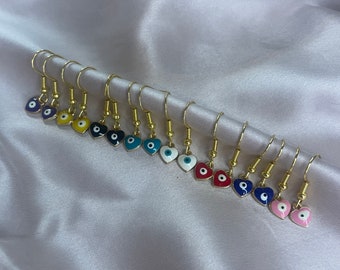 May include: A set of 10 pairs of gold heart-shaped earrings with an evil eye charm. The earrings are available in a variety of colors, including blue, red, yellow, white, and pink.