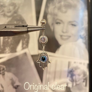 May include: A silver belly button ring with a dangling Hamsa hand charm. The charm features an eye design with blue and black details. The ring also has two clear, round gems. The text "Original clear" is at the bottom.