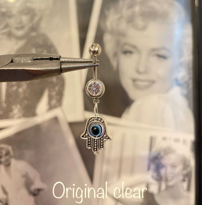 May include: A silver belly button ring with a dangling Hamsa hand charm. The charm features an eye design with blue and black details. The ring also has two clear, round gems. The text "Original clear" is at the bottom.