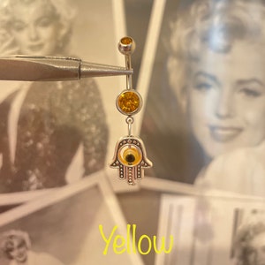 May include: A silver belly button ring with yellow gemstones and a Hamsa hand charm. The ring has a top ball, a middle gemstone, and a dangling Hamsa hand with an eye design. The word "Yellow" is written in a cursive font.
