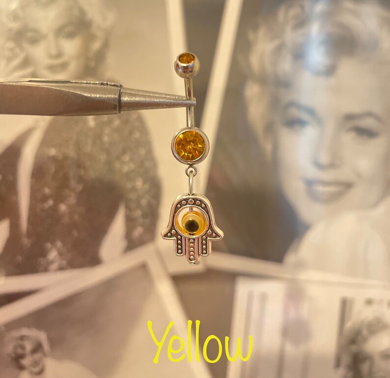 May include: A silver belly button ring with yellow gemstones and a Hamsa hand charm. The ring has a top ball, a middle gemstone, and a dangling Hamsa hand with an eye design. The word "Yellow" is written in a cursive font.