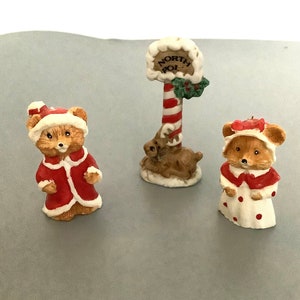May include: Three Christmas figurines: a bear in a red coat and hat, a mouse in a white dress with red polka dots, and a North Pole sign with a reindeer. The sign is red and white striped.