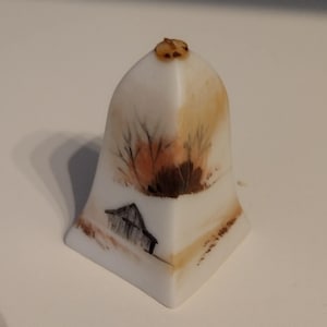 May include: A white ceramic bell with a hand-painted scene of a small house and trees in shades of brown and orange.
