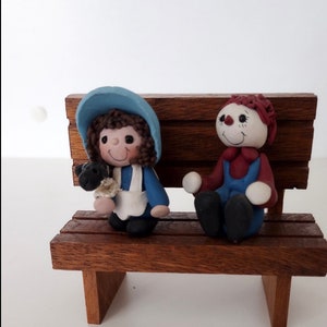 May include: Two handmade figurines of a girl and a boy sitting on a wooden bench. The girl is wearing a blue dress and a blue hat and holding a small black sheep. The boy is wearing a red shirt and blue overalls.