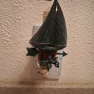 May include: A decorative bronze-colored sailboat-shaped plug-in night light. The night light features a weathervane design with an arrow. The sailboat has a detailed sail and mast. The night light is plugged into a wall outlet.