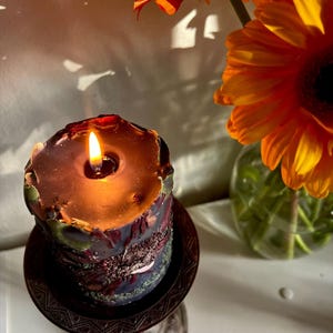 May include: A lit, decorative candle with a dark, textured exterior sits on a detailed, bronze-coloured stand. The candle's wax is a deep brown, and the flame glows brightly. Orange gerbera daisies are in a glass vase in the background.