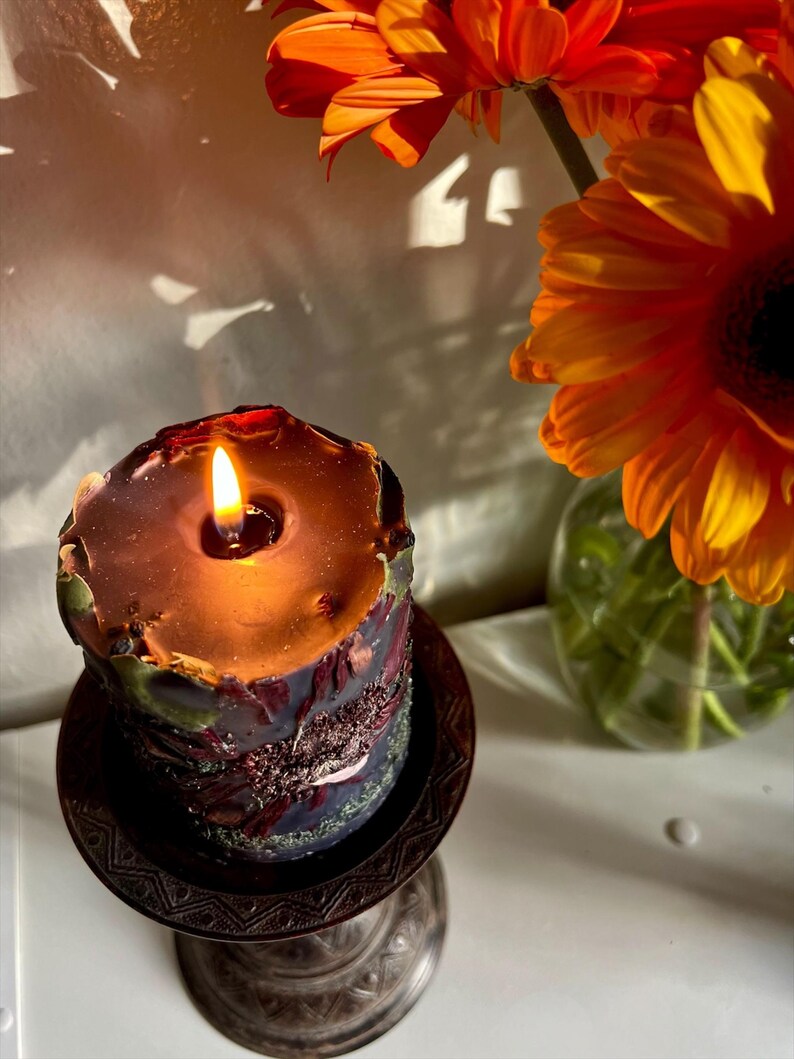 May include: A lit, decorative candle with a dark, textured exterior sits on a detailed, bronze-coloured stand. The candle's wax is a deep brown, and the flame glows brightly. Orange gerbera daisies are in a glass vase in the background.